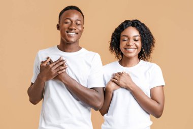 Barışçıl Afro-Amerikalı çift, gözleri kapalı, avuçlarını göğsünde tutuyorlar ve gülümsüyorlar, minnettarlıklarını ve iyi hislerini dile getiriyorlar, sarı stüdyo geçmişi. Mutlu siyah adam ve kadın daha iyisini umuyor.