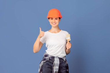 Tamir Servisi Ekibi. Mavi arka planda, Hardhat şapkalı ve boya fırçalı mutlu adam ve kadın, gülümseyen kadın başparmağını kaldırıyor, evi yenilemeye hazır, kopyalama alanı var.