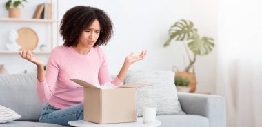 Hayal kırıklığı ve yanlış düzen. Bin yıllık üzgün Afrikalı Amerikalı kadın, içinde alışveriş olan karton kutuya bakar ve gün ışığında oturma odasının içinde kollarını yana doğru açar.