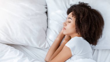 Hafta sonları uyumak, evde boş zaman geçirmek, dinlenmek ve tecrit sırasında dinlenmek. Millennial güzel kıvırcık Afrikalı Amerikalı kadın beyaz yatakta uzanıyor, yatak odasında uyuyor, üst manzara, kopya alanı, profil çizimi