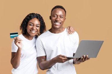 Çevrimiçi bankacılık ve e-ticaret. Mutlu Afro-Amerikalı erkek ve kadın kredi kartı gösteriyor ve sarı stüdyo geçmişi üzerinde çalışıyor. Siyah çift online alışveriş yapıyor, dizüstü bilgisayar kullanarak.