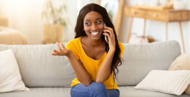 Pozitif genç siyahi bayan cep telefonuyla sohbet ediyor ve evdeki kanepede gülümsüyor. Neşeli Afro-Amerikan kadın kapalı alanda akıllı telefondan biriyle iletişim kuruyor.