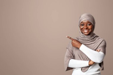 Şuna bak. Tesettürlü Afrikalı Müslüman kadın işaret parmağıyla fotokopi arkaplanındaki boşluğu işaret ederek mesajınız veya tasarımınız için boş yeri gösteren neşeli siyahi kadın.