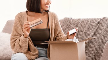 Güzellik kutusu konsepti. Gülümseyen Asyalı kadının portresi teslimat paketini aldı. Makyaj için göz falı paleti tutuyordu. Kozmetik ürünleriyle birlikte karton paketleri boşaltıp evdeki koltukta oturuyordu.