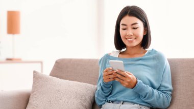 Sosyal Medya Haberleri Kaydırılıyor. Elinde akıllı telefon tutan gülümseyen Asyalı kadının portresi, SMS mesajı yazması, koltukta oturması. İletişim Konsepti. İnternette gezinen kadın, komik videolar izleyen.