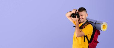 Sırt çantalı ve kameralı neşeli genç gezgin mavi arka planda fotoğraf çekmeye hazır.