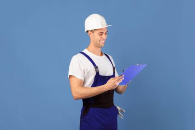 Tulumlar içinde güler yüzlü erkek tamirci ve not defterine sert notlar yazmak, ev geliştirme için bütçe planlayan profesyonel tamirci mavi stüdyo arka planının başında durmak, yer kopyalamak.