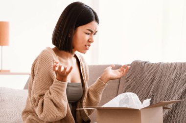 Hasarlı paket. Sinirli Asyalı kadının teslimatını açması, yanlış kutuya bakması, nakliye hatasıyla şok olması oturma odasında kanepede oturması, el sallaması, bedava yer açması.