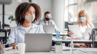 İşteyken fikir üretmek, proje için beyin fırtınası yapmak. Korunaklı maskeli ciddi bir Afrikalı Amerikalı kadın, Coronavirus salgını sırasında modern ofiste çalışan bir pencereye bakıyor.