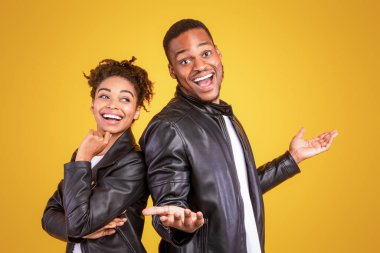 Neşeli Afrikalı Amerikalı erkek arkadaş ve kız arkadaş sırt sırta verip sarı arka planda duruyor, siyah deri ceketler giyiyor. Stüdyo çekimi. Neşe ve Olumlu Duygular Konsepti. Panorama