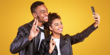 Şık Afrikalı çift telefonda özçekim yapıyor. El hareketleriyle zafer işareti yapıyor. Sarı arka planda duruyor. Panorama, Stüdyo Çekimi