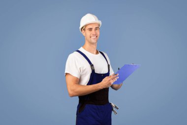 Onarım ve yenileme. Kasklı gülümseyen tamirci, stüdyoda mavi arka planda poz veriyor, panoyu tutuyor, notlar alıyor, pozitif bakım elemanı çalışmaya hazır, kameraya bakıyor.