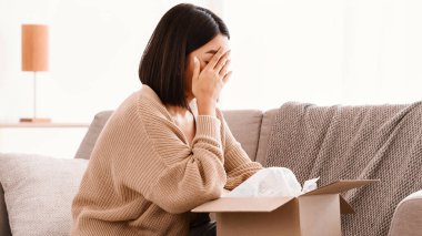 Yanlış paket. Hüsrana uğramış Asyalı kadının kutuları açması, kötü teslimat servisinin evdeki kanepede oturmasından memnun olmaması. Sevkiyat hatası, kopyalama alanı. Yüzünü örten kadın, ağlıyor.