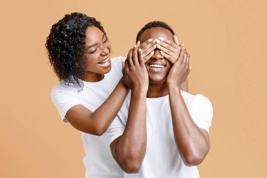 Beautiful black woman covering her man eyes, preparing surprise for birthday over yellow studio background, copy space. Loving wife making surprise for her excited husband on Valentine