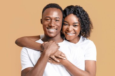 Sarı stüdyo arka planında sarılıp gülümseyen mutlu siyah çift, yakın çekim. Neşeli genç Afrikalı Amerikalı kadın, gülümseyen erkek arkadaşına arkadan sarılıyor, birlikte poz veriyor.