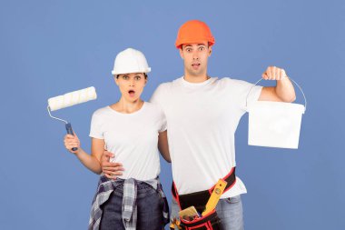 Mavi arka plan üzerinde duran ellerinde boya aletleri olan şok olmuş bir çiftin portresi, boya ve tekerlekli fırçayla kova tutan erkek ve kadın, açık ağızlı kameraya bakıyorlar.