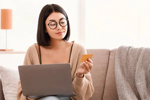 Kolay Ödeme ve İnternetten Alışveriş. Gözlüklü Asyalı kadın internetten sipariş veriyor, dizüstü bilgisayar kullanıyor, kredi kartını tutuyor ve gösteriyor. Bilgisayarlı kadın evdeki koltukta oturuyor.