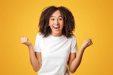 Şans, kazançlar, indirimler ve büyük satışlar. Gülen neşeli, kıvırcık tişörtlü Afro-Amerikalı kadın zaferin duygularını ifade ediyor, sarı arka planda izole edilmiş, boş bir alanda.