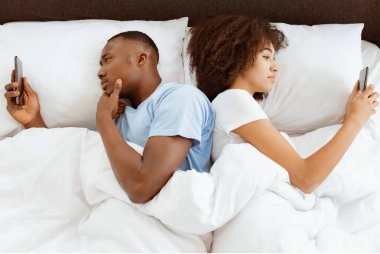 İletişim ve ilişki sorunu. Çift yatakta telefon kullanıyor, sırt sırta uzanıyor, kavgadan sonra görmezden geliyor. Üzgün genç Afro-Amerikan kadın ve adam yatak odasının iç kısmında yazılar yazıyor.