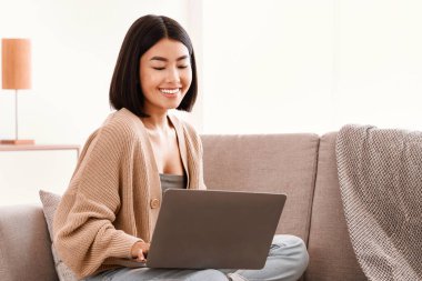 Kanepede oturmuş gülümseyen Asyalı kadın modern dairesinde bilgisayarla oturma odasında çalışıyor. Uzayı kopyala Çalışmak, Eğitim ve Telekomünikasyon İçin Bilgisayar Kullanan Mutlu Bayan Öğrenciler