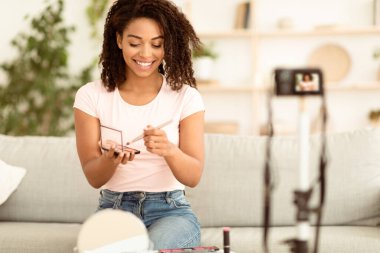 Çevrimiçi Makyaj Dersi. Siyah güzel blogcu kadın internet günlüğü için video hazırlıyor. Evdeki kameranın önündeki koltukta oturan kozmetik ürünlerin nasıl kullanılacağını gösteriyor. Seçici Odaklanma