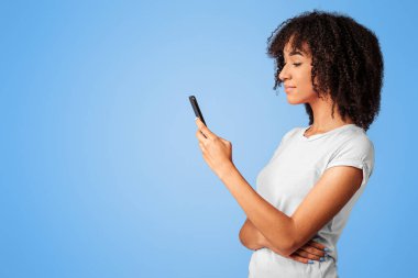 Modern blog öğrencisi ve mobil cihaz çevrimiçi sohbet için. Ciddi bir Afro-Amerikalı kadın, günlük giysiler içinde akıllı telefona bakıyor ve internette sörf yapıyor. Mor arka planda, profilde izole edilmiş.