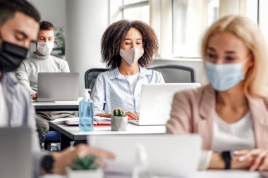 Coronavirus 'ta belgeler ve tartışmalar üzerinde çalış. Koruyucu maskeli adam ve kadın, bürodaki bilgisayarında yazı yazan Afro-Amerikan bayana odaklanıyor, kapatıyorlar, bulanıklaştırıyorlar ve kapatıyorlar.