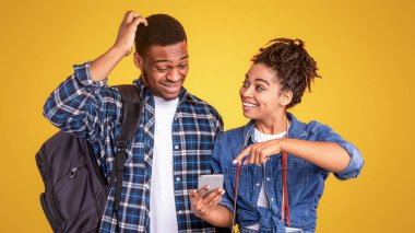 Siyah Kadın, Stüdyoda sarı arka planda durmaya hazırlanan şaşkın arkadaşa telefon gösteren seyahat uygulaması öneriyor. Gezici Uygulama ve Turistler İçin Hizmet Kavramı