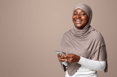 Mobil İletişim. Tesettürlü, dindar, tesettürlü, akıllı telefon pozu veren, cep telefonu tutan, kameraya gülümseyen, yeni uygulamanın keyfini çıkaran, stüdyoda arka planda duran neşeli siyahi kadın.
