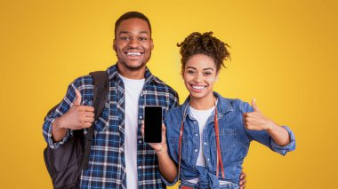 Mutlu Afrikalı Gezginler Çift Akıllı Telefon Boş Ekran Hareketlerini Gösteriyor, Gezici Mobil Seyahat Uygulamasını Sarı Stüdyo Arkaplanında Tavsiye Ediyor. Turist Başvurusu Kavramı Onaylanıyor