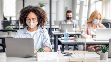 Virüs salgını sırasında birlikte çalışmak için modern cihazlar. Koruyucu maskeli Afro-Amerikalı bir kadın bilgisayarında akıllı telefon, antiseptik ve koruyucu camlar ve boş bir alanla yazı yazıyor.