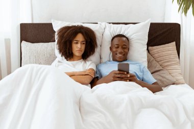 Modern ilişkiler, iletişim ve aile sorunları. Memnuniyetsiz binlerce yıllık Afro-Amerikan kadın elinde akıllı telefonuyla gülümseyen kocanın telefonuna bakar.