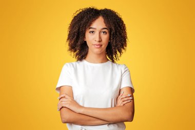 Zeki öğrenci ve insani duygular. Kendine güvenen, kıvırcık saçlı, kolları çapraz beyaz tişörtlü, sarı arka planda izole edilmiş, stüdyo çekimi olan Afro-Amerikan bir kadın.
