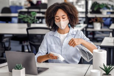 Koronavirüs ve yeni normallere karşı sağlığını korumak için işyeri hijyeni. Maskeli Afrikalı Amerikalı kadın ofis içinde masa ve laptopu dezenfekte etmek için sprey ve kağıt havlu kullanıyor.