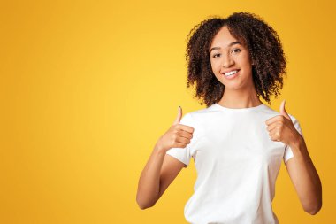 Doğal insan duyguları ve blog yazarından harika bir teklif. Gülümseyen bin yıllık Afro-Amerikan kız öğrenci beyaz tişörtlü kıvırcık saçlı baş parmaklarını gösteren, sarı arka planda izole edilmiş, boş bir alan.