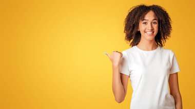 İnsanlar, promosyon, duygular, tanıtım, reklam ve danışmanlık. Beyaz tişörtlü gülümseyen Afro-Amerikan kadın, sarı arka planda, panoramada, stüdyo çekimlerinde izole edilmiş, boş yeri gösteriyor.