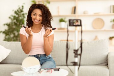 Güzellik Blogu. Afro-Amerikalı Kadın Blog Yazıcı Makyaj Ürünleri İncelemesi Evdeki Kameranın Yakınındaki Koltukta Oturan Video Yapıyor. Makyaj Blog Kavramı. Seçici Odaklanma