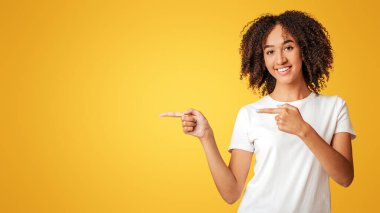Reklam ve sunum konsepti. Gülümseyen, binlerce yıllık Afro-Amerikan kadını beyaz tişörtlü, kıvırcık saçlı, sarı arka planda izole edilmiş boş bir yeri gösteren.