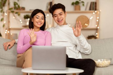 Neşeli Çinli çift dizüstü bilgisayarlarında video görüşmesi yapıyor evdeki koltukta oturuyor, el sallıyor. Evli Eşler Uzaktan Arkadaşları Arıyor. Uzak İletişim Konsepti