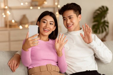 Japon çift evdeki koltukta oturan akıllı telefona cep telefonuyla el sallayarak video görüşmesi yapıyor. Cep telefonu kavramı için Uzaktan İletişim, Çevrimiçi Video Konferansı Uygulaması