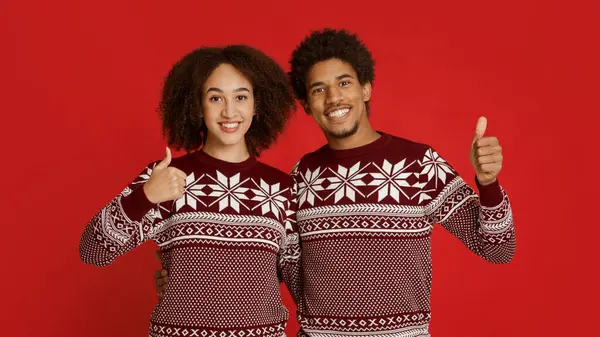 Mutlu ve olumlu, mükemmel ve onay işareti. Mutlu binlerce yıllık Afro-Amerikan aile çifti Noel süveterleri içinde kırmızı arka planda, panoramada, boş alanda, stüdyo çekimlerinde başparmak gösteriyorlar.
