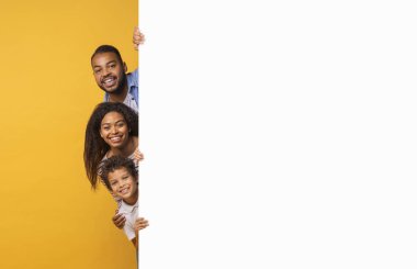 Beyaz reklam panosunun arkasından bakan üç kişilik neşeli siyah aile, mutlu bir ebeveyn ve genç oğul sarı arka plana karşı duruyorlar, yeni bir teklif öneriyorlar, kopyalama alanı.