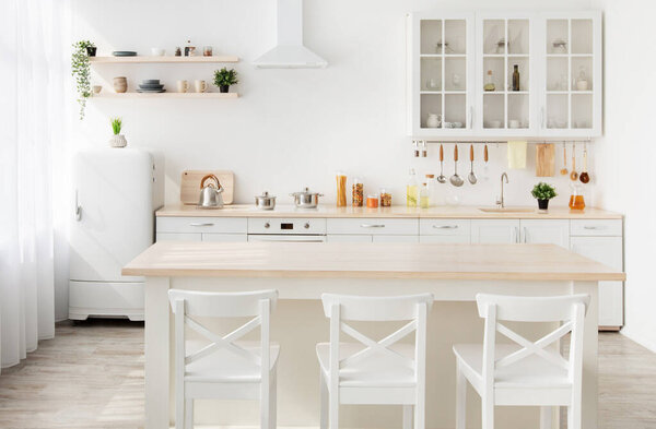 Стиль минимализма. Стильная кухня из белого и коричневого дерева. Обеденный стол, посуда и кухонные принадлежности на полках, маленький белый холодильник у окна