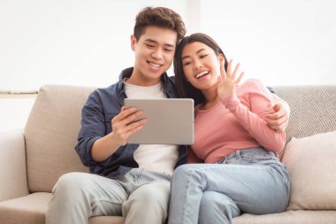 Çinli bir çift dijital tabletle video görüşmesi yapıyor, evdeki koltukta oturup uzaktan iletişim kuruyor, bilgisayar ekranına el sallıyor. Uzaklık İletişim Konsepti. Boşluğu Kopyala