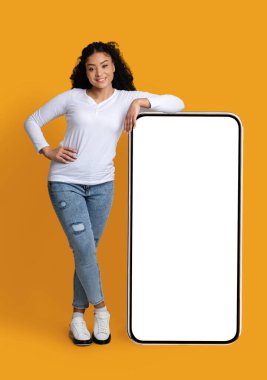 Cool Mobil Uygulama. Neşeli siyahi kadın, beyaz ekranlı büyük akıllı telefona yaslanıyor, tasarımın veya reklamın için kopyalama alanı gösteriyor, sarı arka planda duruyor, görüntüyü taklit ediyor.