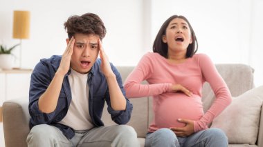 Ani Doğum sancısı. Hamile Asyalı Kadın Dokunaklı Karnı Ağrılı Spazm Geçirirken Şok Olan Koca Evde Koltukta Otururken. Hamilelik ve Doğum Sorunları