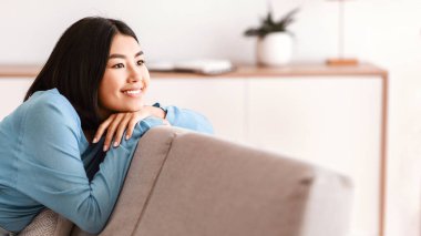 Hafta sonu ve Boş Zaman Konsepti. Gülümseyen Asyalı bir kadının portresi. Kanepede dinleniyor. Kendine güveni tam mutlu bir bayan. Kanepeye uzanıyor, boş zamanların tadını çıkarıyor ve tatilin hayalini kuruyor.