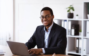 Ofiste modern dizüstü bilgisayarla çalışan neşeli Afro-Amerikan iş adamı, kopyalama alanı. Takım elbiseli ve gözlüklü mutlu genç siyah yönetici online çalışıyor, e-posta yazıyor ya da müşterilerle sohbet ediyor.