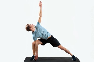 Yoga pozisyonunda duran güçlü genç adam stüdyo arka planında spor minderinin üzerinde duruyor, uzun bir portre. Atletik milenyum sporcusu esneme egzersizleri yapıyor, pilates yapıyor. Refah kavramı