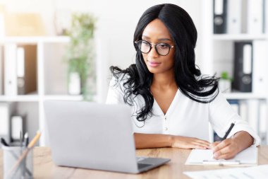 Ofiste dizüstü bilgisayarda çalışan mutlu zenci iş kadını. Odaklanmış Afrikalı Amerikalı kadın çevrimiçi iş toplantısı veya webinar sırasında not alıyor, önemli bilgiler yazıyor